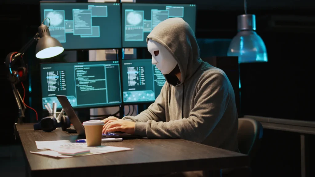 Person wearing a hoodie and mask working on a laptop with multiple screens displaying code.